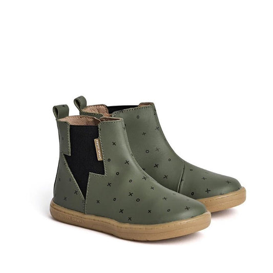 The Pretty Brave Electric Boot Khaki XO features olive green leather, black elastic side panels, tan soles, pull tabs, and playful black X patterns—perfect for kids wanting standout style.