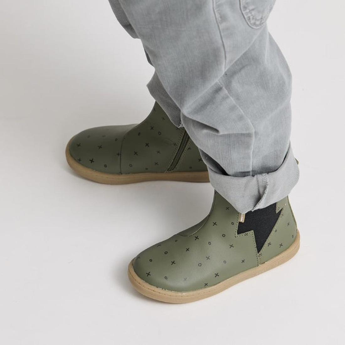 A child wears light gray pants and Pretty Brave Electric Boot Khaki XO kids leather boots with small black X patterns and a black lightning bolt design on the sides, standing on a white background., 28805, 28806, 9420075305335, 9420075305342, 942007530535
