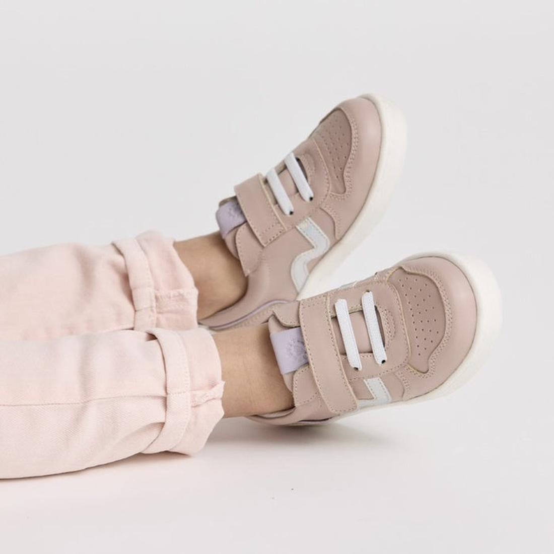 A child wears light pink pants and Pretty Brave XO Trainer Blush sneakers—blush pink with white soles and Velcro straps—while lying or sitting with legs extended on a white background., 28764, 9420075304024, 9420075304031, 9420075304048, 9420075304055, 94