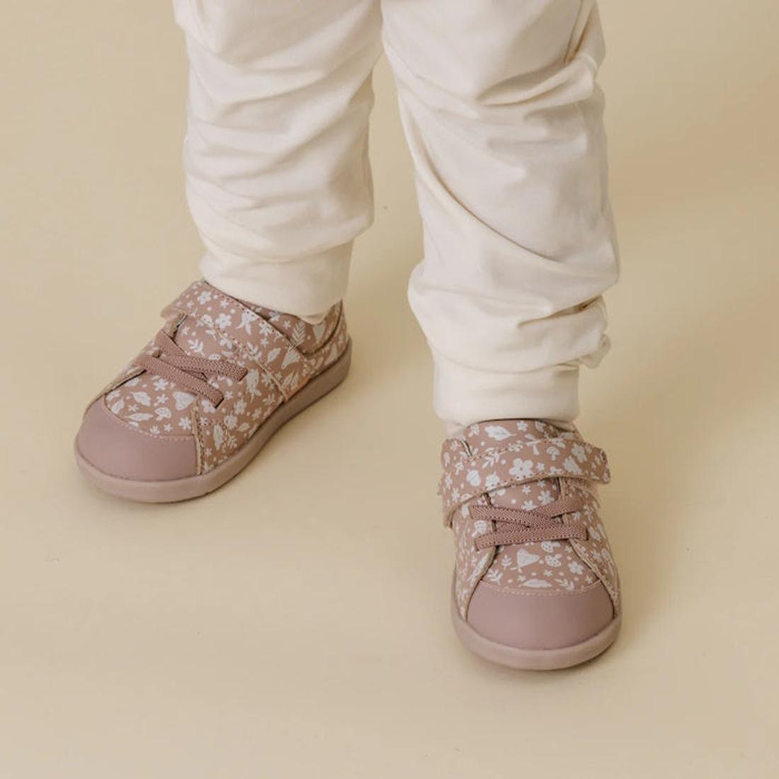 Child wearing pink Boston floral first walker sneakers with white pants on a beige background.,
9420075347922,
9420075347915,, 9420075347922, 9420075347915, 46563