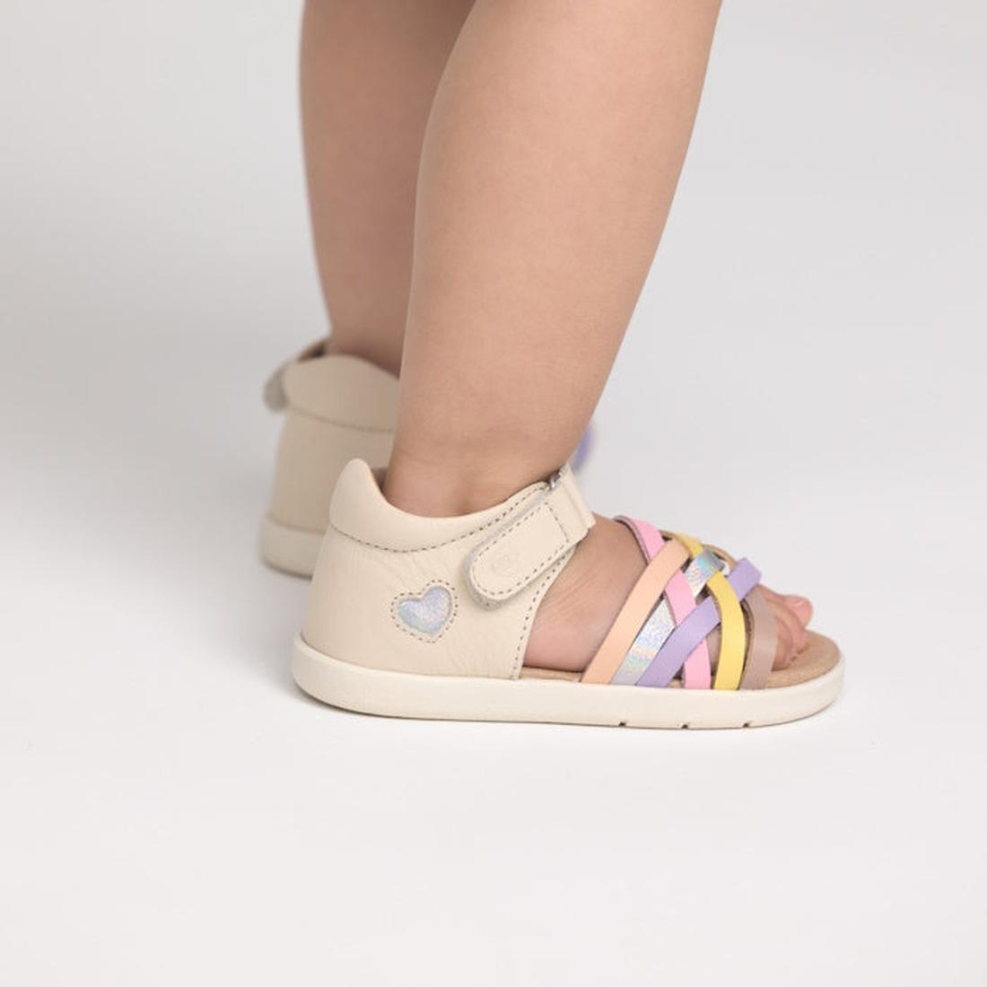 A toddler stands on a white surface wearing Pretty Brave 1st Walker Eden Rainbow Leather Open Toe Sandals, featuring podiatrist-approved design, pastel pink, yellow, purple, and silver straps, a blue heart detail, and an adjustable strap., 9420075355705, 