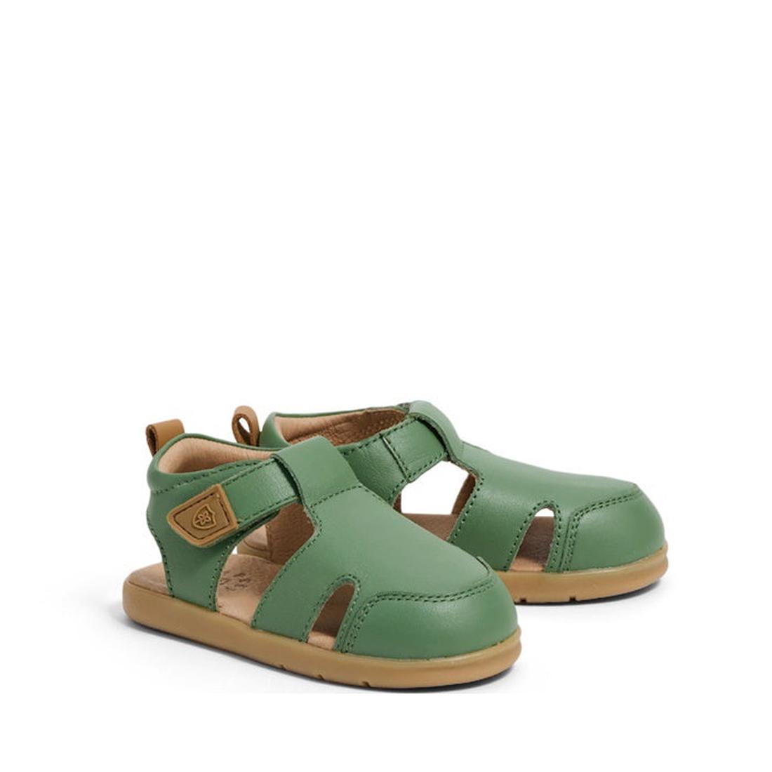 The Pretty Brave 1st Walker Eli Grass Leather Closed Toe Sandal is a green and tan children's sandal with cut-out sides, an adjustable Velcro strap, and flat soles, displayed on a plain white background.