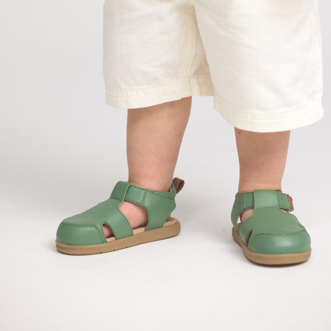 A young child stands on a light surface, showing only their legs and feet in loose white pants and Pretty Brave 1st Walker Eli Grass Leather Closed Toe Sandals with adjustable Velcro straps, podiatrist-approved for little walkers., 9420075358669, 94200753