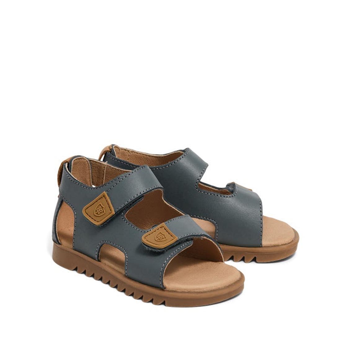 The Pretty Brave Child Finley Indigo Leather Open Toe Sandal features supportive, indigo leather straps, tan hook-and-loop fasteners, cushioned tan footbeds, and brown ridged rubber outsoles. Sandals are shown from a slight angle.