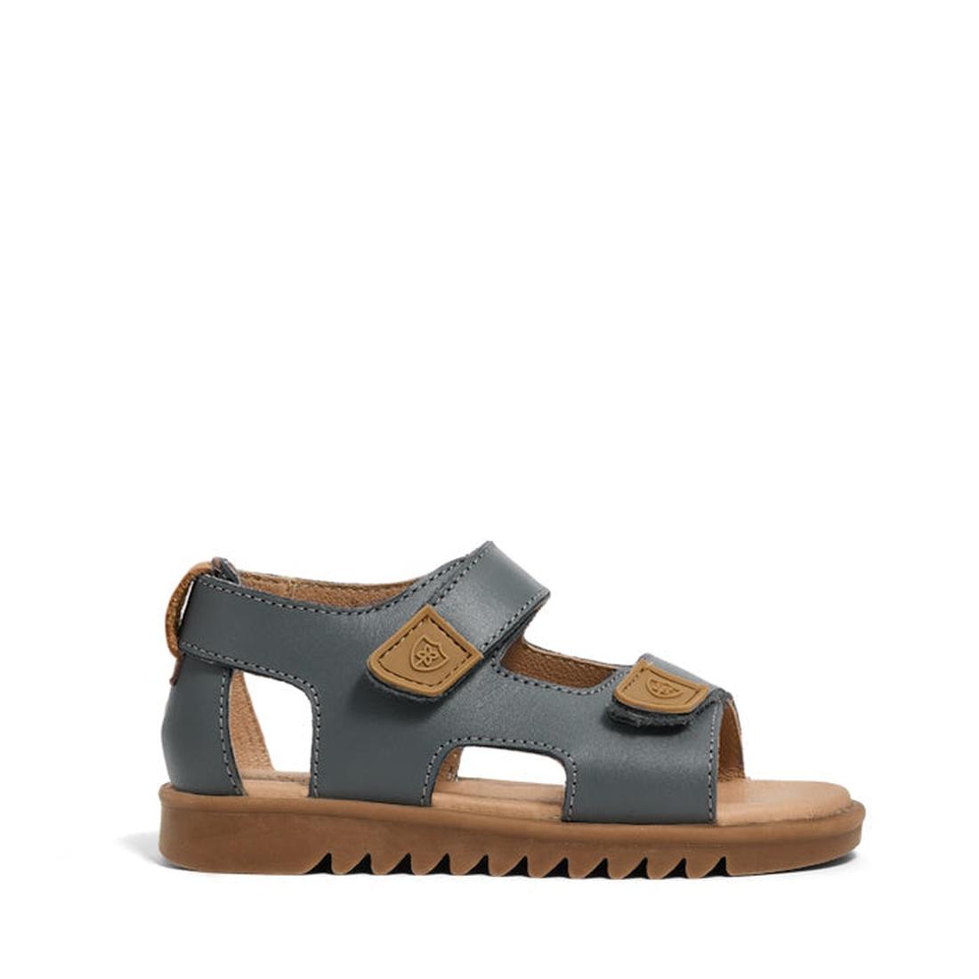The Pretty Brave Child Finley Indigo Leather Open Toe Sandal features a gray leather upper, brown adjustable Velcro straps, open toe and heel, cut-out sides, cushioned footbed, and a brown ridged rubber sole. Shown in profile on white background., 9420075