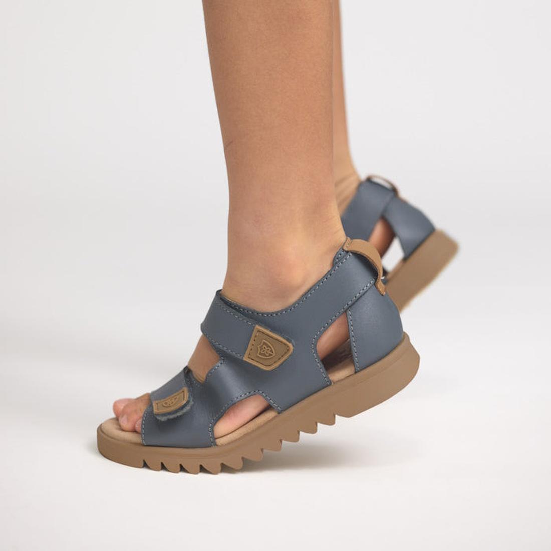 A child stands on one foot against a plain white background, wearing Pretty Brave Child Finley Indigo Leather Open Toe Sandals featuring adjustable Velcro straps, thick ridged soles, and a cushioned footbed., 9420075358478, 9420075358461, 9420075358454, 9
