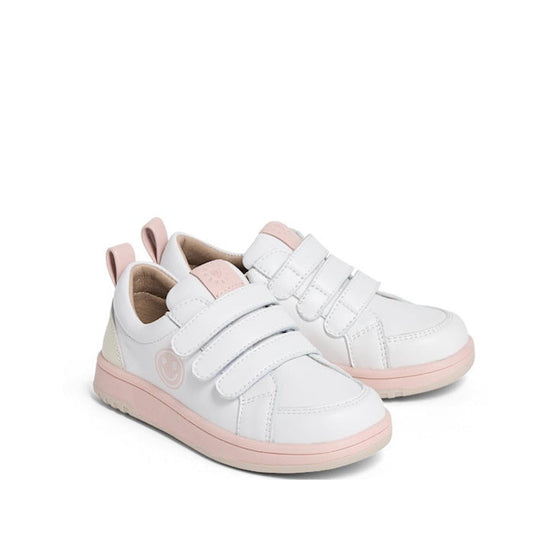 The Pretty Brave Happy Trainer White/Blush Sneaker is a kids’ leather shoe in white with light pink soles, toe caps, and accents. It features two velcro straps, a heel pull tab, and a small smiley face on the side for added charm.