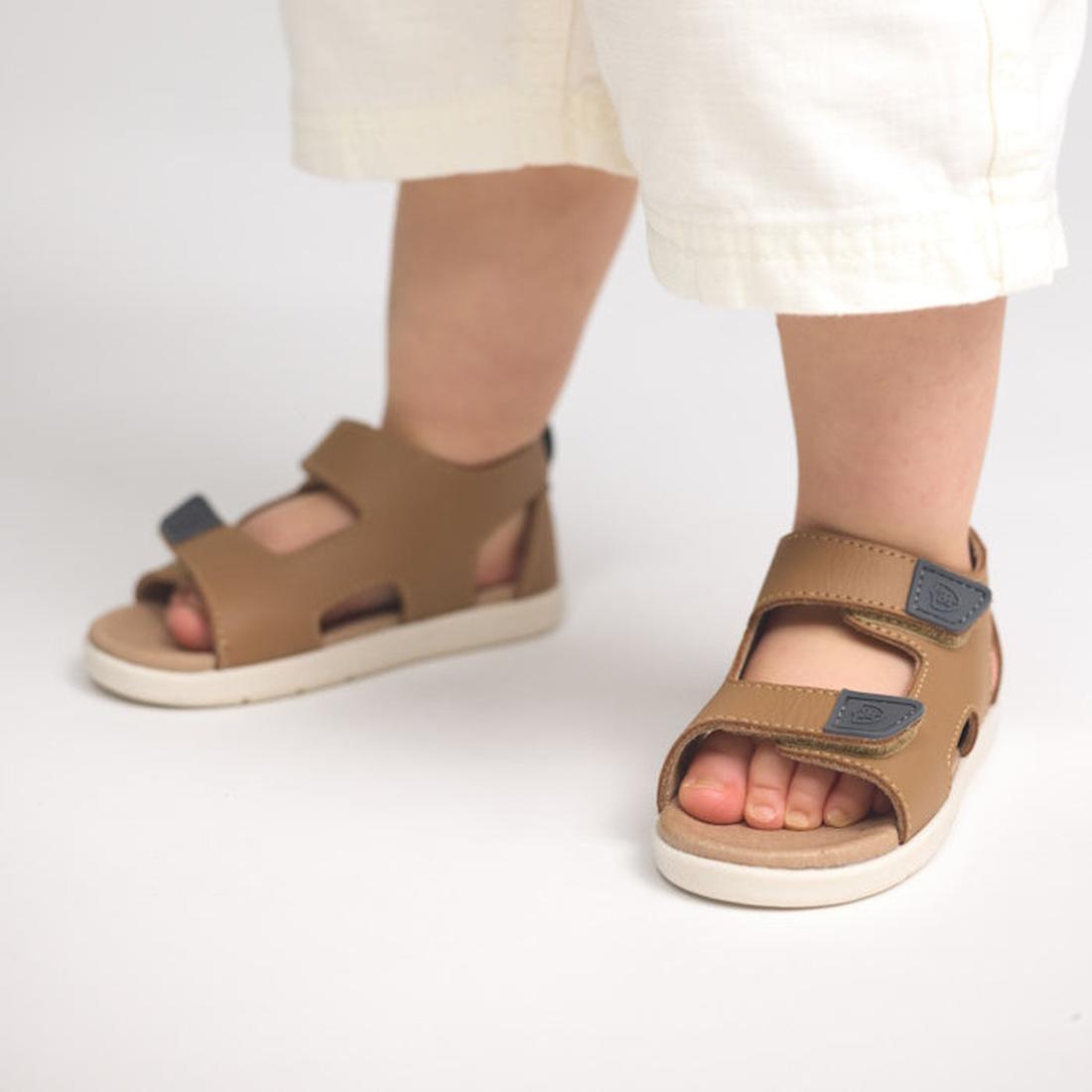 A child stands on a light-colored surface wearing white pants and Pretty Brave 1st Walker Jack Tan Leather Open Toe Sandals with adjustable Velcro straps., 9420075358713, 9420075358706, 9420075358690, 9420075358683