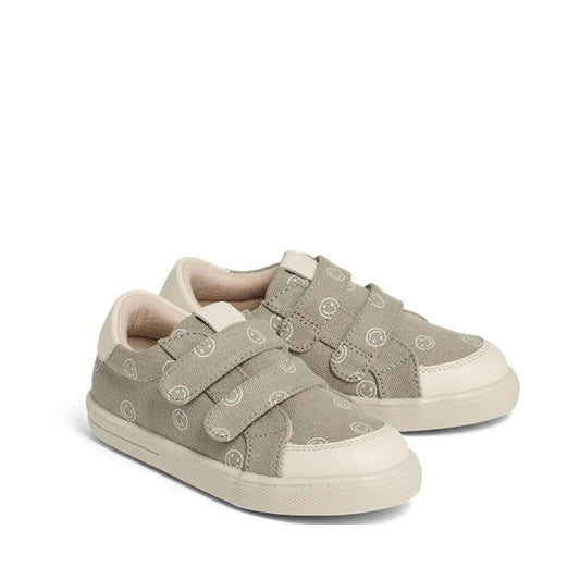 A pair of Pretty Brave Otto Organic Canvas Trainers in khaki smiley, featuring a leather toe buffer, white soles, two Velcro straps, subtle circular pattern on natural cotton canvas, and a soft beige lining. Podiatrist recommended.