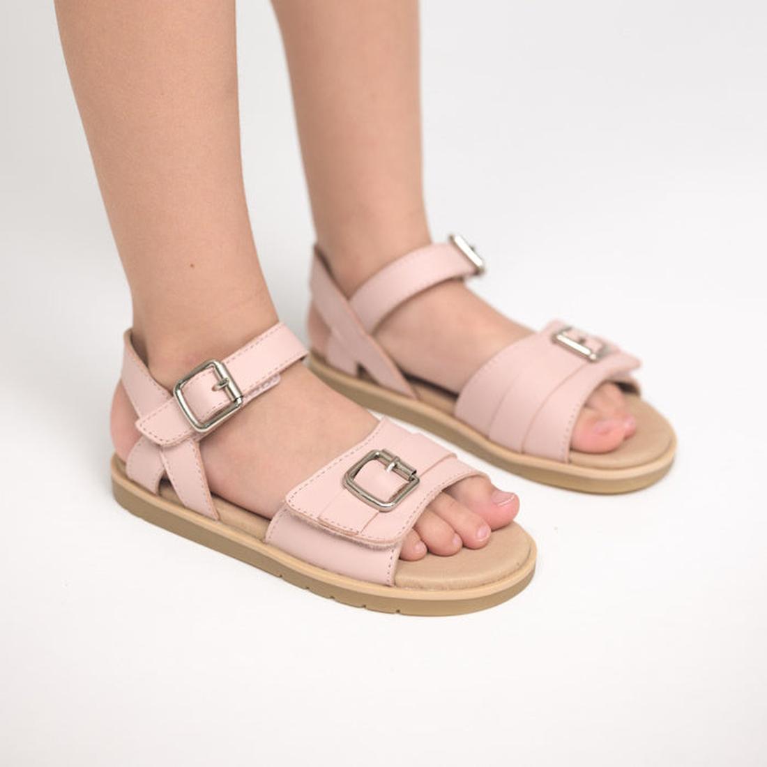 A child stands on a white surface wearing Pretty Brave Child Piper Soft Pink Leather Open Toe Sandals, featuring large silver buckles, a leather footbed, and straps over the toes and around the ankle., 47173, 9420075357938, 9420075357921, 9420075357914, 9