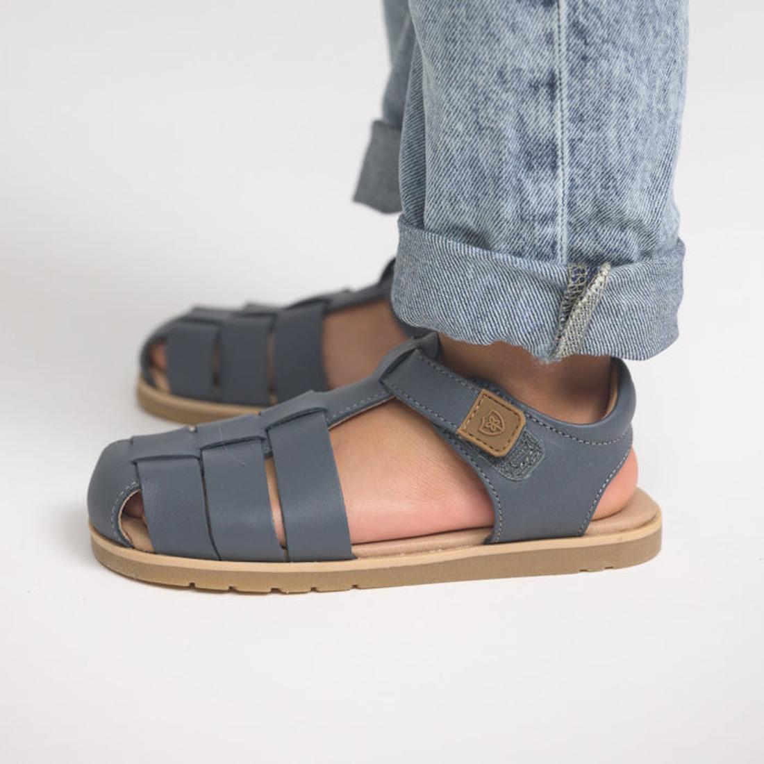 A child wearing rolled-up light blue jeans and Pretty Brave Child Rocco Indigo Leather Closed Toe Sandals with tan soles, standing on a light surface., 9420075357556, 9420075357549, 9420075357532, 9420075357525, 9420075357518, 9420075357501, 9420075357495