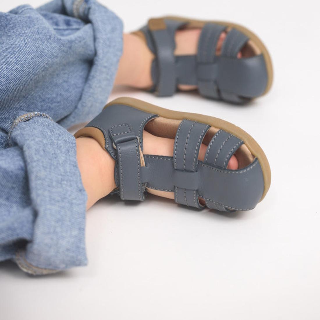 A child sits on a white surface wearing cuffed blue jeans and Pretty Brave 1st Walker Theo Indigo Leather Closed Toe Sandals with tan soles, highlighting the supportive design and focusing on the sandal and lower legs., 9420075358768, 9420075358751, 94200