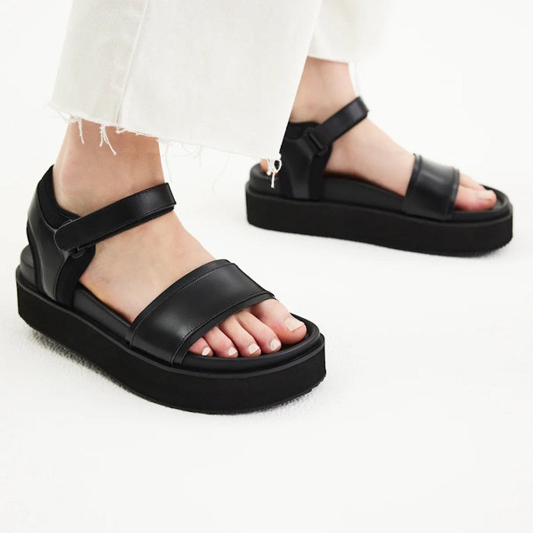 A person wearing white, frayed-hem pants and Rollie Womens Ace Sandal All Black Leather with thick, lightweight platform soles and two wide straps, standing on a light-colored surface., 9339554333878, 9339554333885, 9339554333892, 9339554333908, 933955433