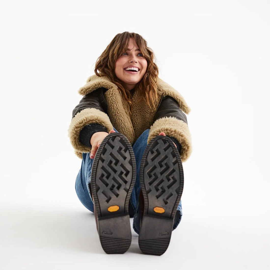 A smiling woman in a shearling jacket and jeans sits on the floor, legs extended to show the Vibram outsole of her Rollie Womens Chelsea Rise Vintage Mocha Leather Boots, against a plain white background., 9339554341682, 9339554341699, 9339554341705, 9339
