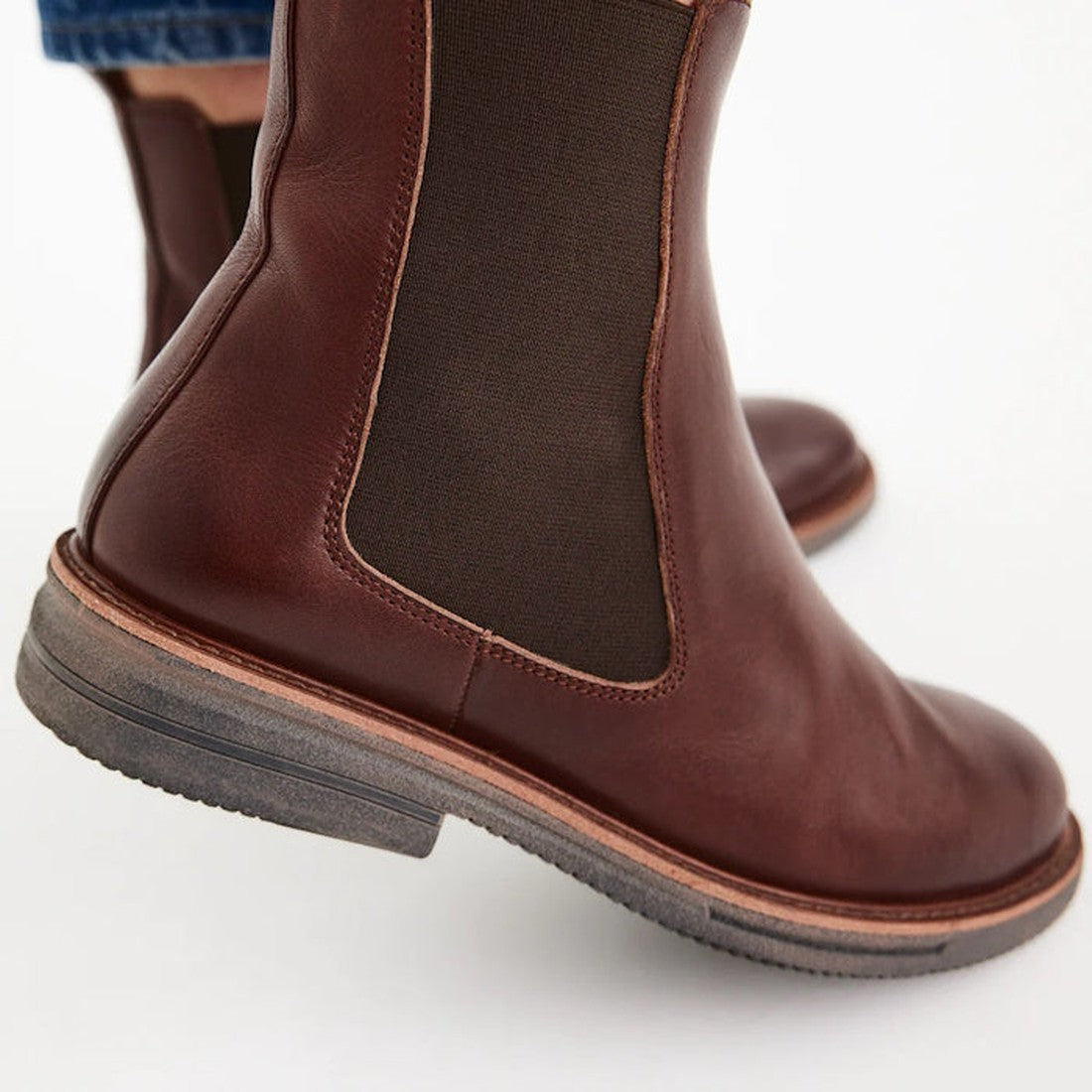 Close-up of a person wearing Rollie Womens Chelsea Rise Vintage Mocha Leather Boot with dark elastic side panels, light brown soles, and a Vibram outsole, standing on a white background., 9339554341682, 9339554341699, 9339554341705, 9339554341712, 9339554