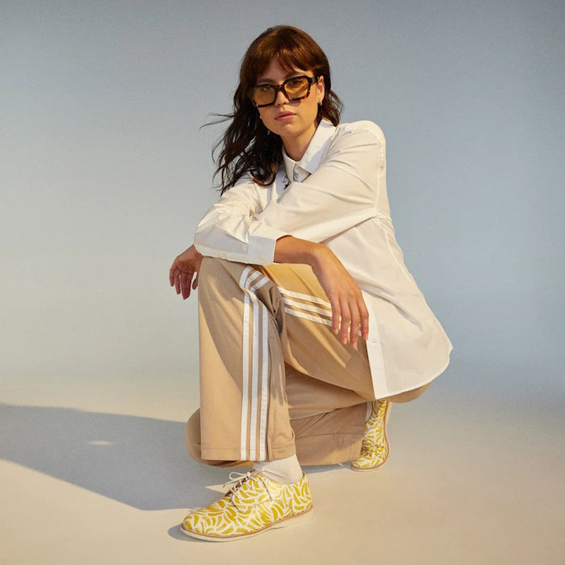 Squatting against a plain, softly lit background, a person wears sunglasses, a white button-up shirt, tan pants with white stripes, and the Rollie Womens Derby Sunshine Splash Leather Shoe for sneaker-like comfort., 9781760774783, 9781760774790, 978176077