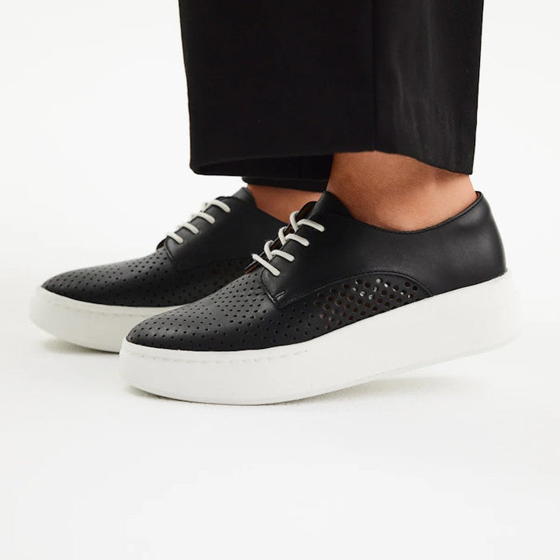 A person wearing Rollie Womens Derby City Punch Black Leather Trainers with thick white platform soles and black pants stands on a white background. These sneakers are orthotic-friendly with a removable footbed for comfort., 9339554259628, 9339554259635, 