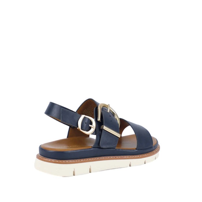 The Soultbush Garie Navy Blue Womens Sandal with Back Strap features a cushioned footbed, tan sole, gold buckle, and crisscross straps. Shown from the side and slightly behind on a white background, this sandal is perfect for effortless coastal living.