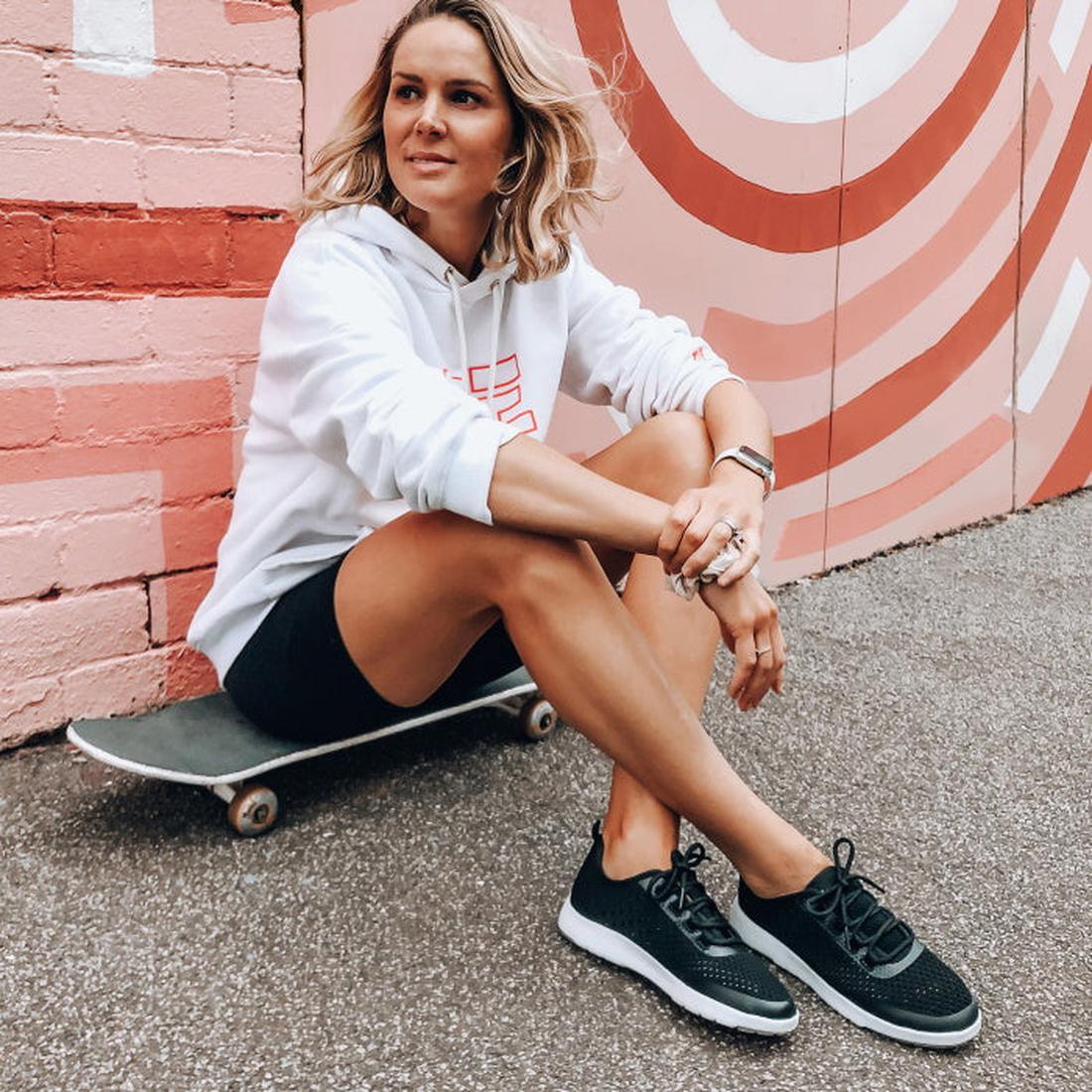 A young woman in a white hoodie and black shorts sits on a skateboard, wearing comfy EMU NZ Miki Black Lace-Up Sneakers with soft soles.  ksnz