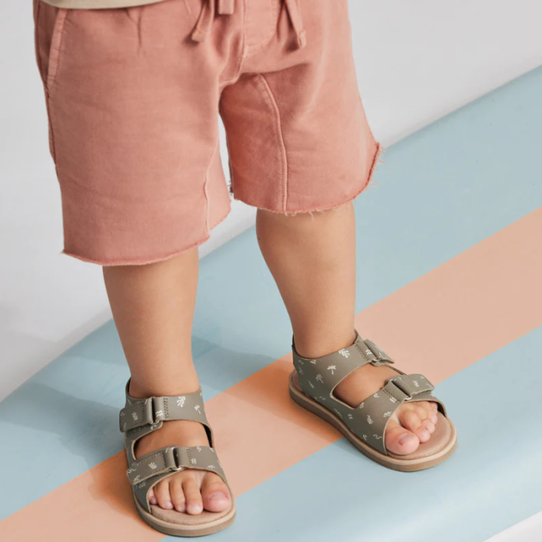 A young child in light pink shorts and Pretty Brave Wilder Reef Green Kids Sandals (sizes 29-31, not returnable), featuring white patterns and adjustable velcro straps, stands on a pastel blue and peach striped surface., 9421036359503, 9421036359510, 9421