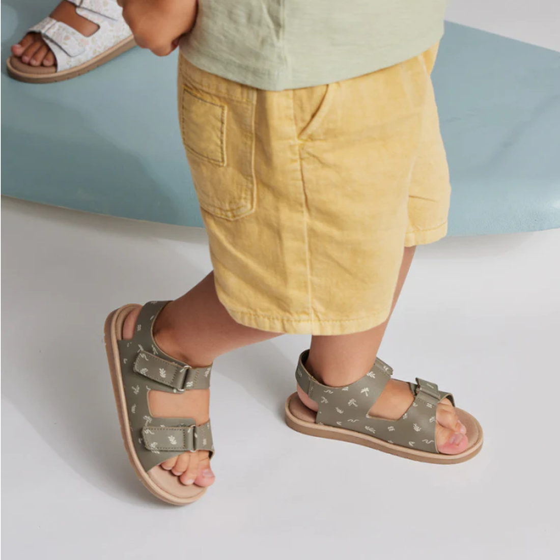 A young child in yellow shorts wears Pretty Brave Wilder Reef Green Kids Sandals (29-31, NOT RETURNABLE) with adjustable velcro straps on a light blue surface. Another child's feet in white kids sandals are visible in the background., 9421036359503, 94210