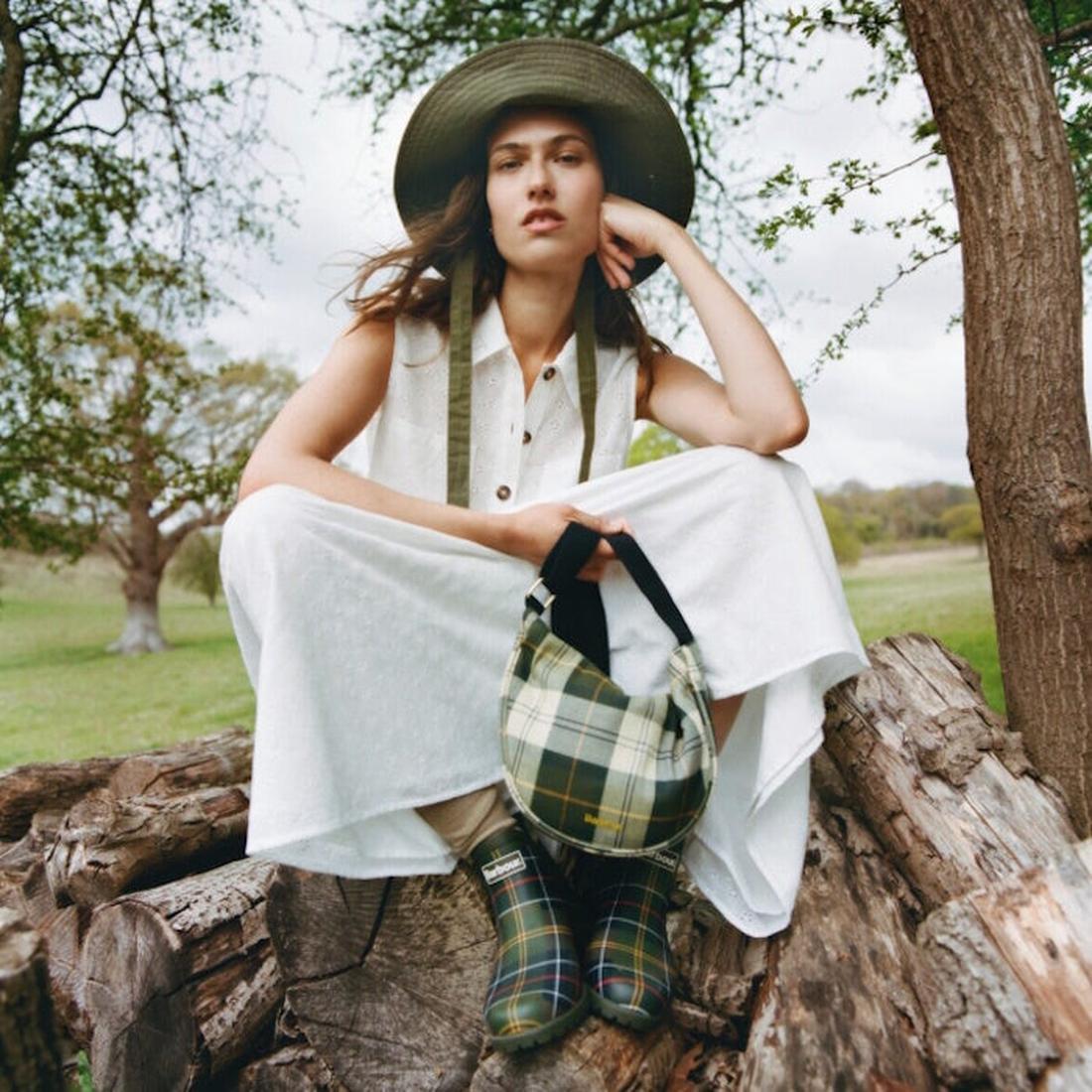 A woman in a white dress and wide-brimmed hat sits on a large log outdoors, holding a plaid bag and wearing Barbour Wilton Classic Tartan Chelsea Wellingtons Womens Rainboot, with trees and grass in the background., 194972145934, 194972146030, 19497214612