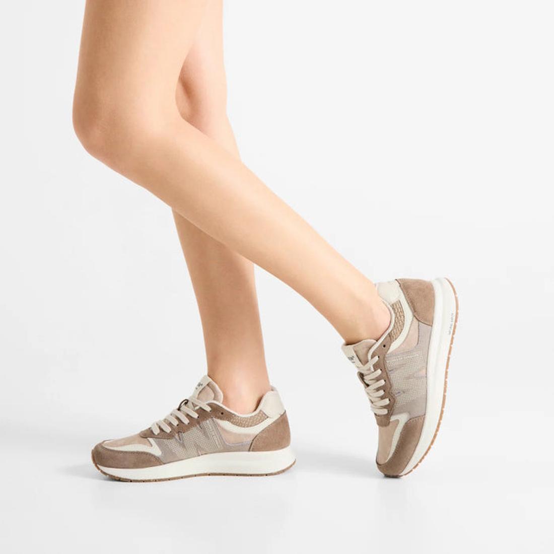 A person with bare legs wears Woden Rigmor Heritage Coffee Cream Lace Up Sneakers featuring beige laces and LWG certified suede, standing on one leg with the other slightly bent against a plain white background., 50259, 5713326695113, 5713326695120, 57133