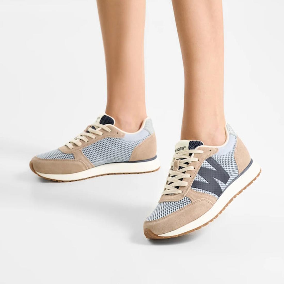 A person stands on a white background, showing only their lower legs and feet in Woden Ronja Icon Moonstone Multi Lace Up Sneakers featuring light blue, beige, and gray colors with recycled mesh panels, white laces, and gum soles., 50270, 5713326699678, 5