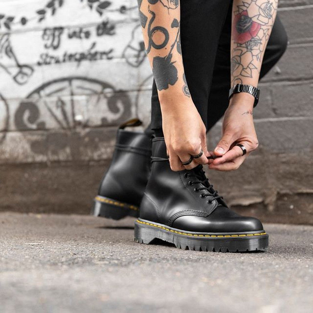 A person with tattooed arms laces up Dr Martens - Bex 1460 8 Up Black Leather Boots, featuring yellow stitching and a chunky Bex sole, while standing on a street by a graffiti-covered wall., 190665298758, 190665298765, 190665298772, 190665298789, 19066529