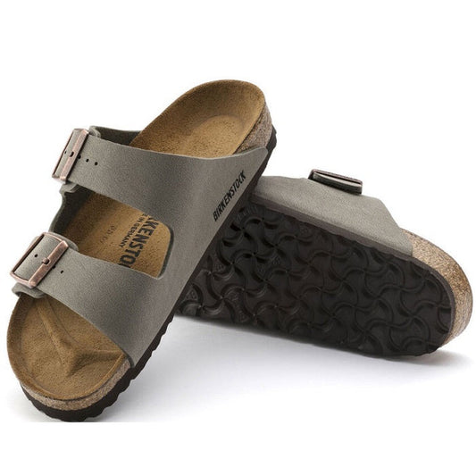 A pair of Birkenstock Arizona Birkibuc Stone Narrow Width sandals with two adjustable buckles, a contoured cork-latex footbed, and one sandal on its side highlighting the textured sole.