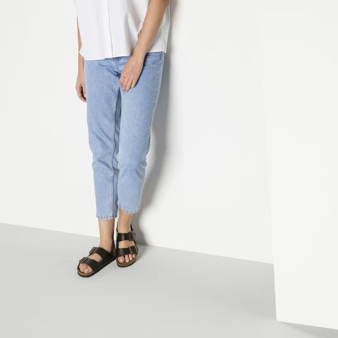 A modela wearing light blue denim 3/4 pants and a white shirt standing against a wall wearing Birkenstock Arizona leather sandal slides in dark bown narrow fit. showing double straps across the top of the foot.,4013871078616,4013871018407,4013871019466,40, 4013871045779, 4013871074724, 4013871058250, 4013871059127, 4013871019558, birb43, birb44, birb45, birb46