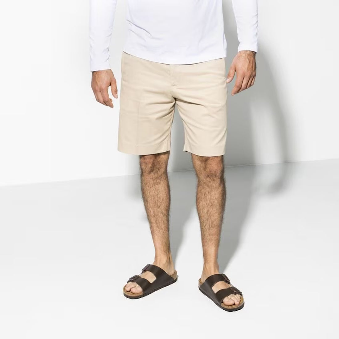 A man, face unseen, stands against a white background wearing a white long-sleeve shirt, beige shorts, and BIRKENSTOCK Arizona Smooth Leather Dark Brown Regular Width sandals with a cork-latex footbed.,
4013871066453,
4013871069898,
4013871076551,
4013871