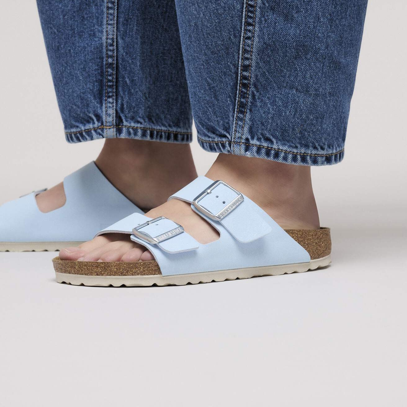 Close-up of a person wearing Birkenstock Arizona Finished Sky Blue Birkibuc – Regular Fit sandals with silver buckles and a cork-latex footbed, paired with cropped blue jeans, standing on a light surface., 4067707550128, 4067707550166, 4067707550203, 4067