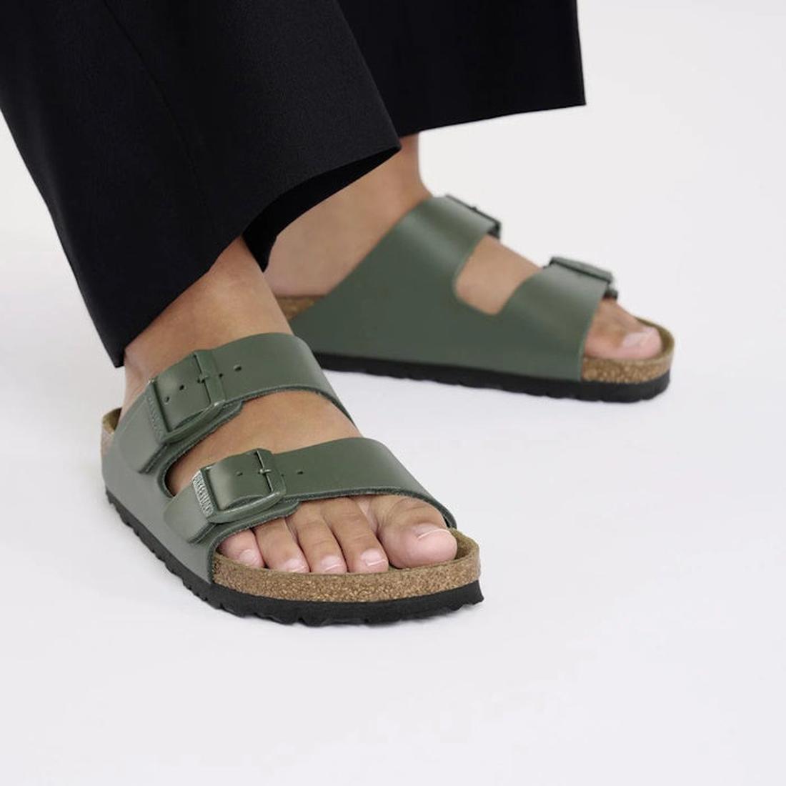 A person wearing Birkenstock Arizona Smooth Leather Thyme sandals (regular width) with black pants, standing on a white surface., 45282, 45283, 45284, 4066648927563, 4066648927600, 4066648927648, 4066648927686, 4066648927723, 45290, 45291, 45292, 45293