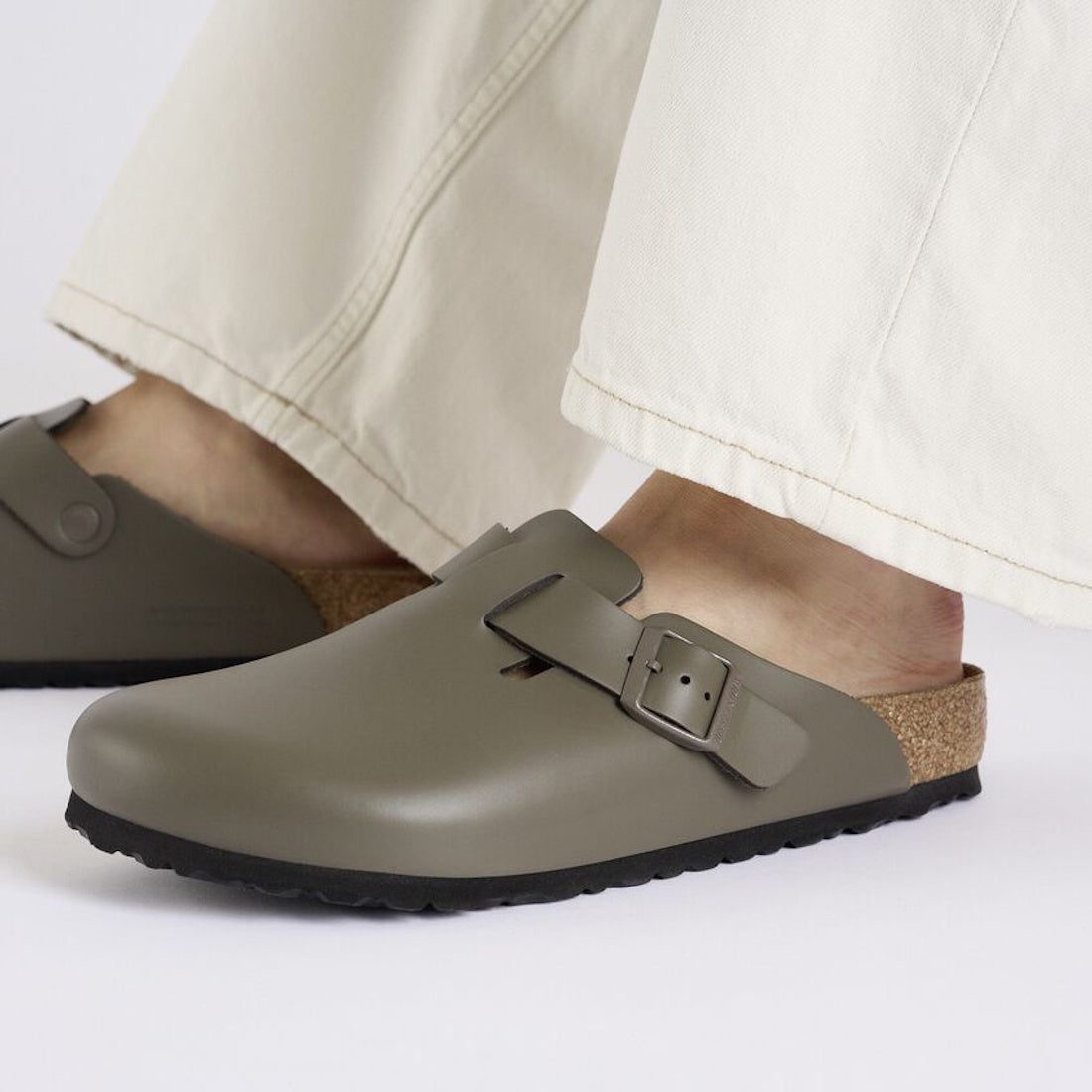 A person wearing cream-colored pants and Birkenstock Boston Clog Smooth Leather Regular in Concrete Grey Dark, featuring a contoured cork-latex footbed and buckle strap, stands on a white background., 4066648924166, 4066648924203, 4066648924241, 406664892, 4066648924289, 4066648924326, 4066648924364, 4066648924401, 4066648924449, 4066648924487
