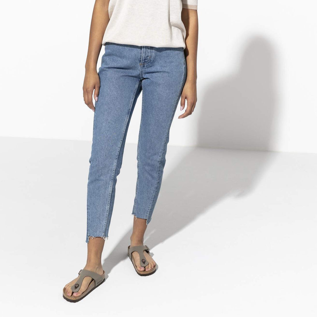 A person stands against a white background wearing a white short-sleeve top, blue skinny jeans with frayed hems, and Birkenstock Gizeh BirkiBuc Stone Grey sandals (regular width). Their head and upper torso are not visible., 48753, 48754, 4013871810414, 4