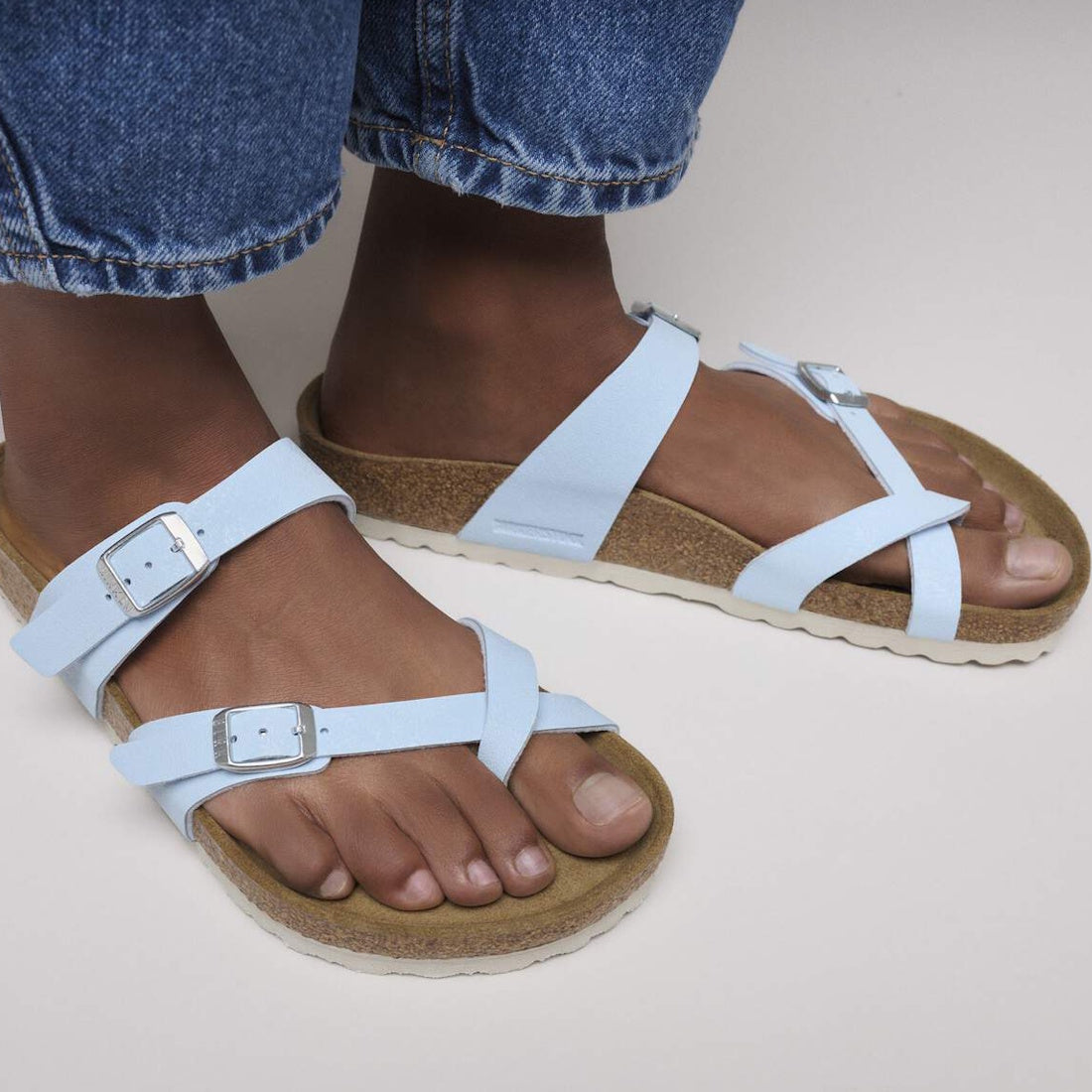 Close-up of a person wearing Birkenstock Mayari Birko Flor Birkibuc Finished Sky Blue Regular Width sandals with silver buckles and cork soles, paired with rolled-up blue jeans against a plain light background.,4067707274901,
4067707274949,
4067707274987,, 4067707275021