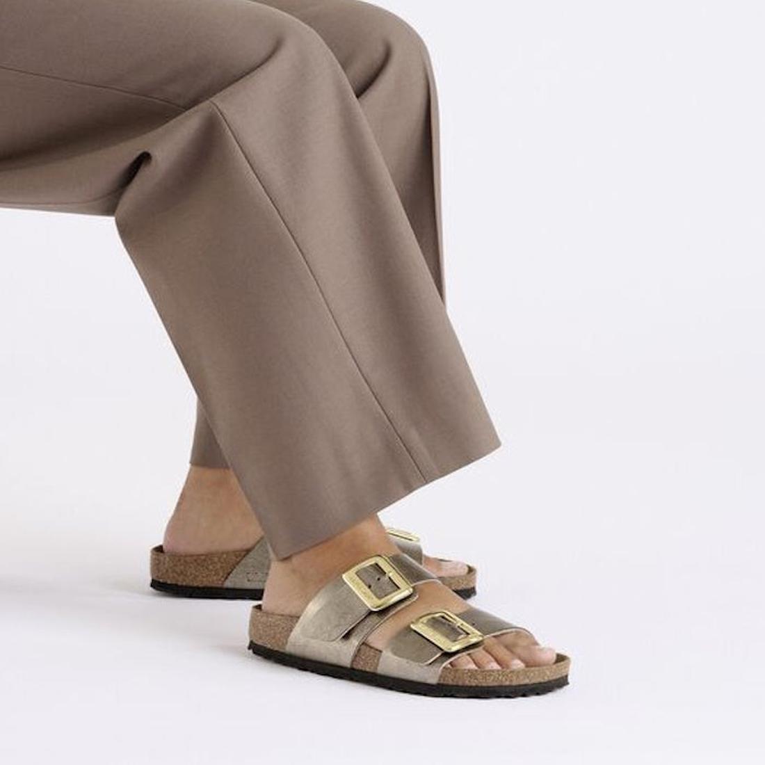 Person wearing brown sandals with gold buckles and beige pants on a light gray background,
4066648998228,
4066648998266,
4066648998303,, 45674, 4066648998181, 4066648998228, 4066648998266, 4066648998303, 4066648998341