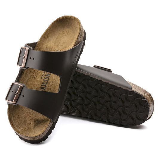 A pair of BIRKENSTOCK Arizona Smooth Leather sandals in dark brown (regular width) with two adjustable buckle straps and a cork-latex footbed. One sandal stands upright as the other leans to display the textured sole.