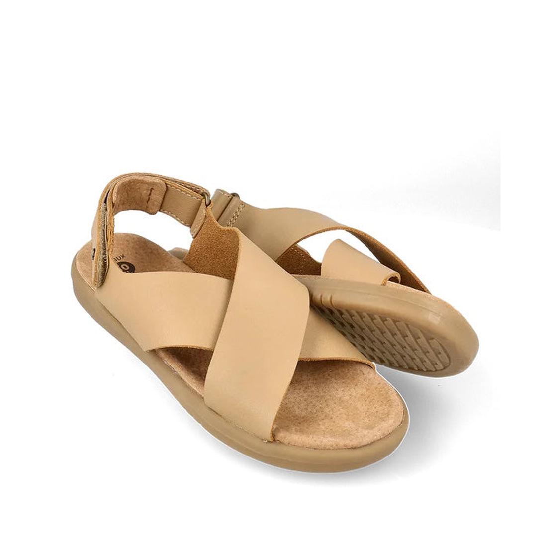 A pair of Bobux Kid+ Plus Glide Parchment Beige Open Toe Sandals with thick crisscross leather straps, adjustable heel strap, and textured rubber sole; one stands upright while the other leans to show the sole.