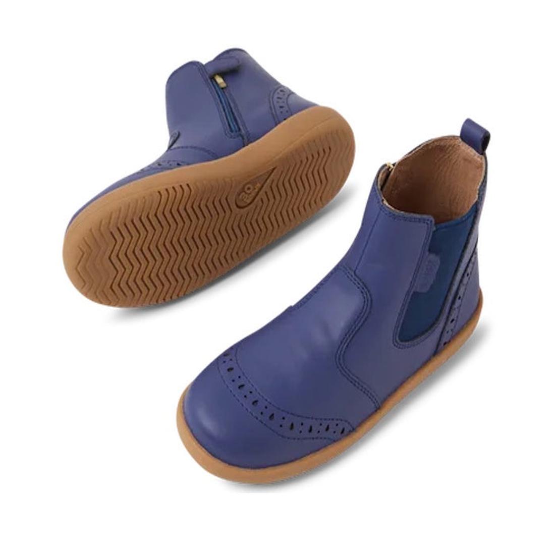 The Bobux Kid+ Billie Leather Boot Future Dusk Blue/Purple 852002 is a unisex kids’ boot featuring blue leather, tan rubber soles, side elastic panels, perforated details, and a textured sole. One boot stands upright while the other lies on its side.
