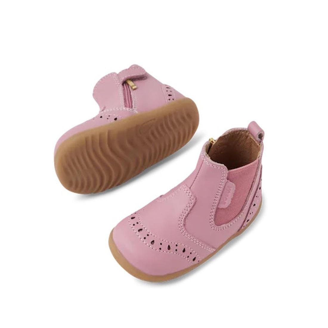 The Bobux Step Up Billie Girls Boot Lilas Pink 752003 is a toddler ankle boot in pink leather with brown rubber soles, side zippers, and decorative perforations, shown on a white background.