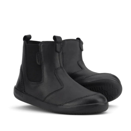 The Bobux Kid+ Black School Leather Boot Jodphur Black Sole Shoes feature classic Jodhpur-inspired design with black leather, elastic side panels, front and back pull tabs, and textured rubber soles. One boot stands upright, the other reveals its sole.