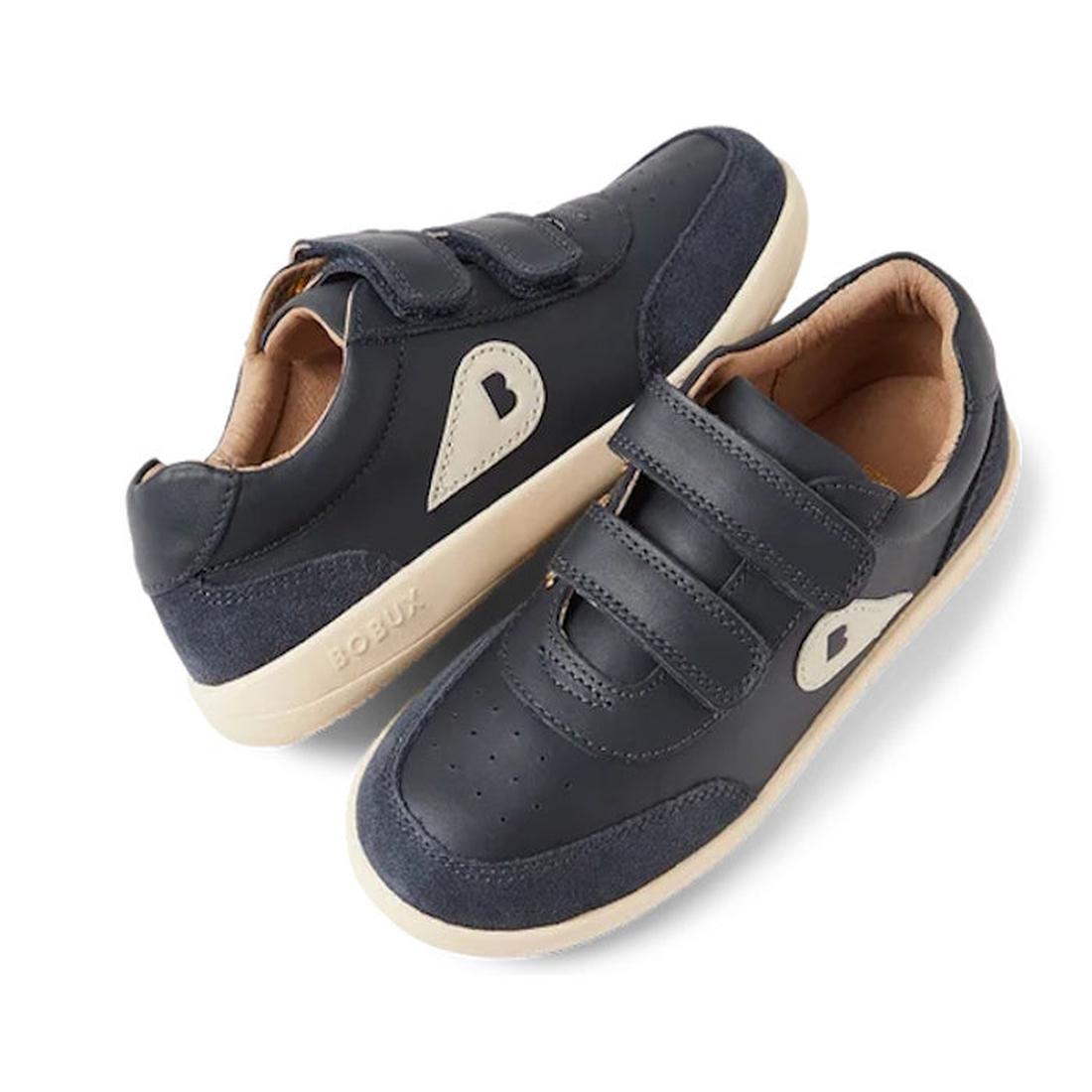 The Bobux Kids Plus KP Champ Sneaker Navy 850509 is a pair of navy blue leather kids’ sneakers with beige soles, beige accents, two velcro straps, a soft tan lining, ankle support, and a heart-shaped side detail.