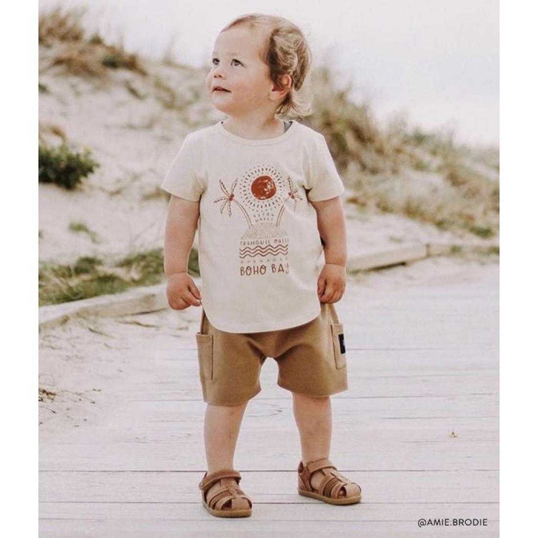 A young child stands on sandy ground, wearing a cream boho tee, tan shorts, and Bobux Step Up Todders Sandal Roam 729204B in Caramel. The child smiles and looks upward with greenery and sand in the background., 15014, 9420062081464, 9420062081471, 9420062