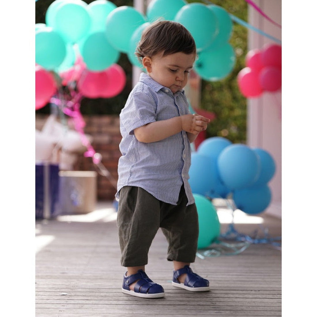 A young boy in a blue striped shirt, dark pants, and Bobux Step Up US Tidal Navy Closed Toe Sandal 732503 stands on a wooden floor with colorful balloons and party decorations in the background., 9420068502574, 9420068502581, 9420068502598, 9420068502604
