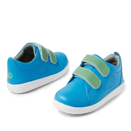 The Bobux KP Kid Plus Grass Court Brilliant Blue 841012 kids’ shoes feature bright blue uppers, white soles, green Velcro straps, and heart-shaped perforations on the sides. The trainers are displayed side by side on a white background.