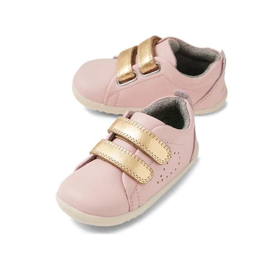 The Bobux Step Up Grass Court Trainer Ballet Pink 734218 is a light pink leather toddler shoe with two metallic gold Velcro straps, white soles, and a soft gray inner lining, shown on a white background.