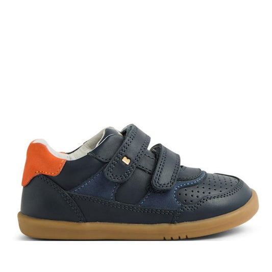The Bobux IW Iwalk Hunter Leather Sneaker in navy features a tan rubber sole, orange heel accent, perforated toe, and two hook-and-loop straps. Displayed from the side on a white background.