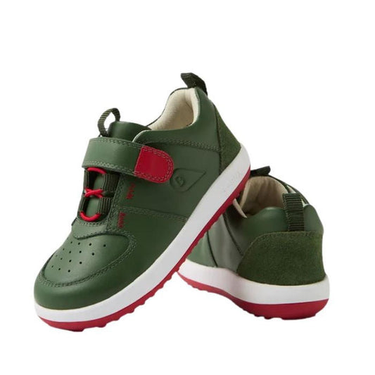A pair of Bobux IW Iwalk Backspin Leather Boots in bronze green with red accents, white soles, hook and loop straps, and red elastic laces. One boot stands upright while the other leans against it, displaying side and back views.